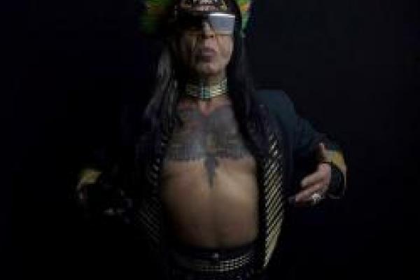 Dark skinned person with a black jacket open to reveal a tattooed chest with sunglasses and a colorful headpiece