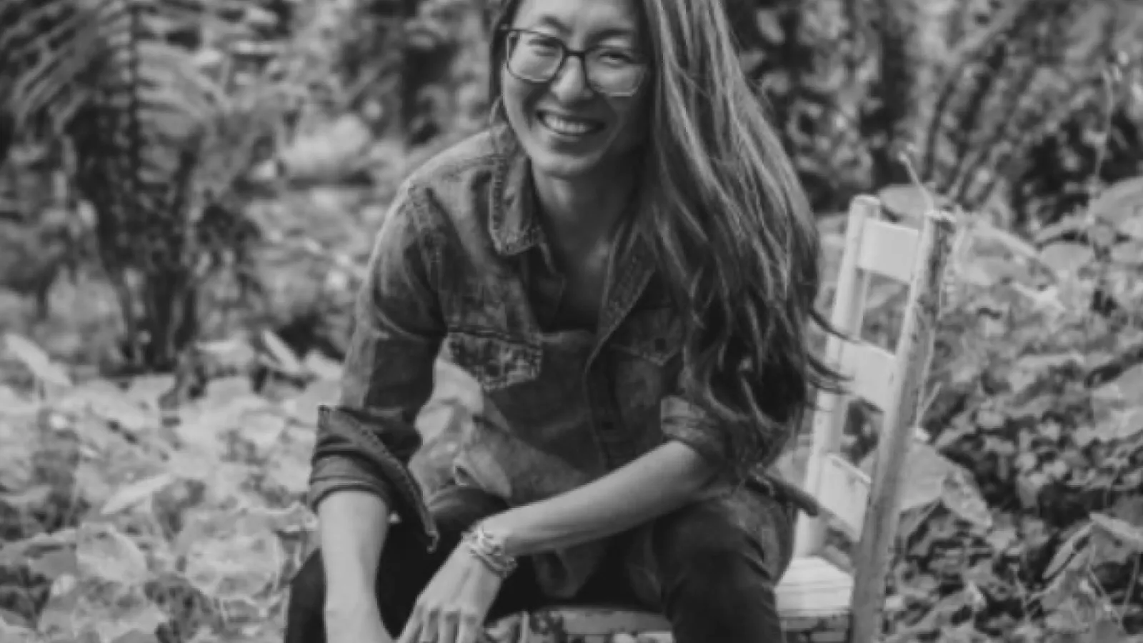 Black and white image of Mia Mingus sitting on a wooden chair in a jungle setting