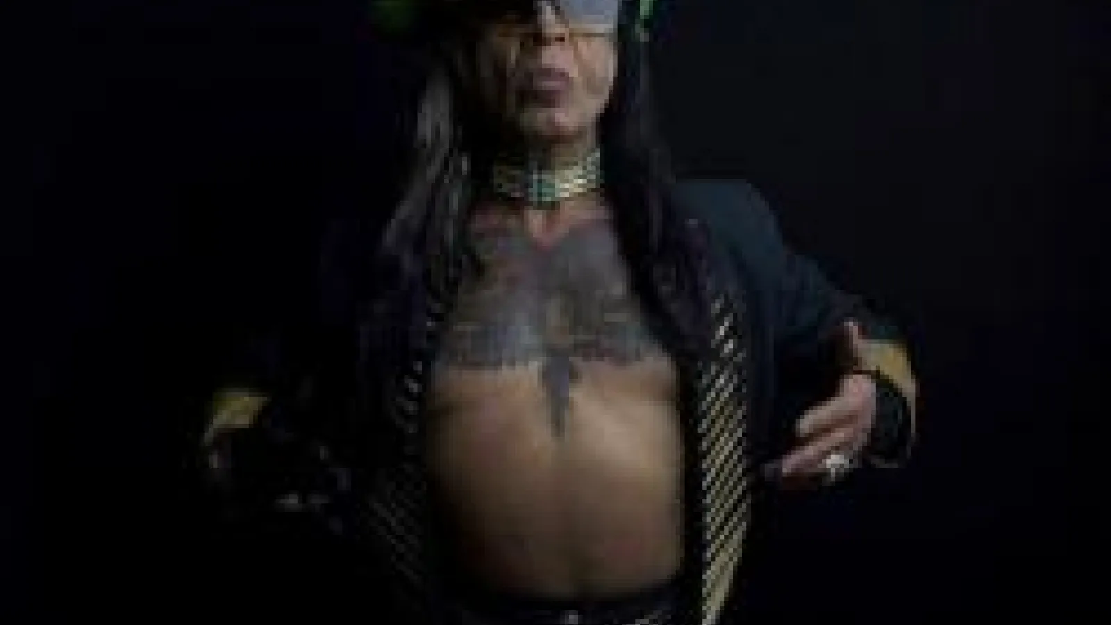 Dark skinned person with a black jacket open to reveal a tattooed chest with sunglasses and a colorful headpiece