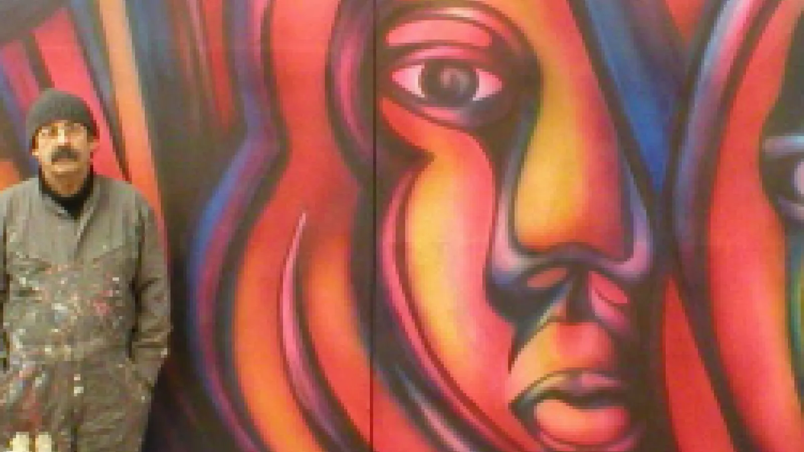 Mono Gonzalez standing to the left of a large orange and red painted mural of a woman's face