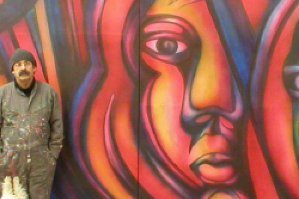 Mono Gonzalez standing to the left of a large orange and red painted mural of a woman's face Mono Gonzalez standing to the left of a large orange and red painted mural of a woman's face
