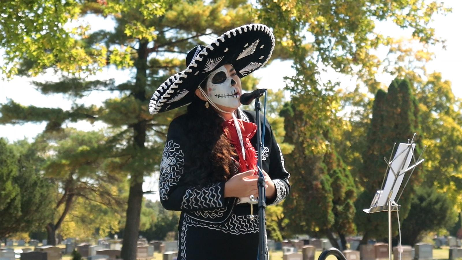A woman in a mariachi costume and skull makeup singing into a microphone